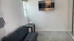 A modern living room with a black sofa, a ceiling fan, a small table with a tissue box, and a large flat-screen TV mounted on the wall.