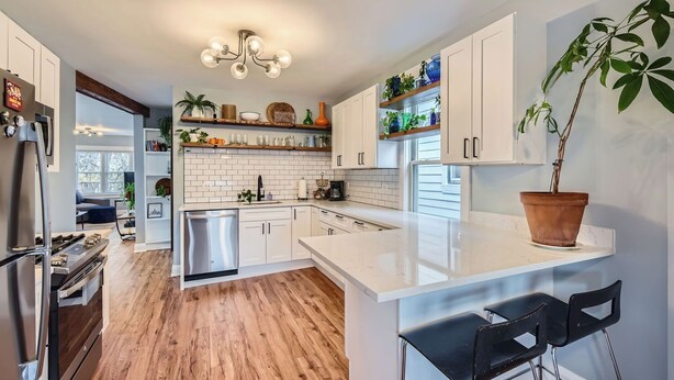 A modern kitchen with white cabinets, a stainless steel refrigerator, a dishwasher, and a wooden floor. There is a potted plant on the counter and a hanging light fixture above the sink.