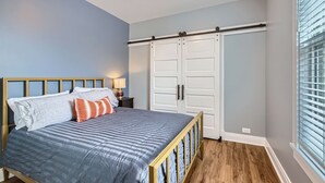 A bedroom with a bed, bedside table, lamp, wardrobe, and a window with blinds.