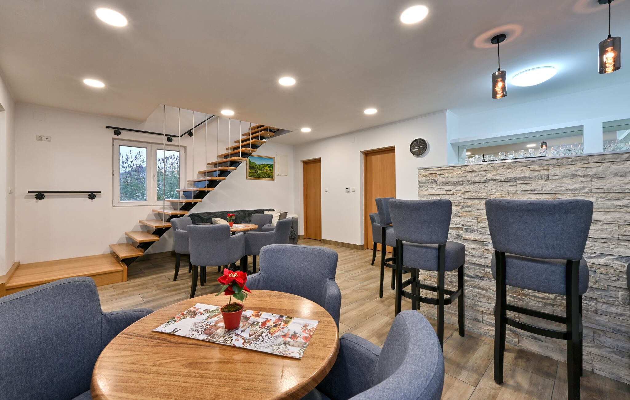 A modern living room with a wooden staircase, grey upholstered chairs, a round wooden coffee table with a red flower, and a stone accent wall.