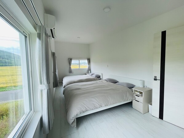 A modern bedroom with two beds, a window with curtains, a bedside table with a lamp, and a view of a green landscape. - TOMATO CUBE (Asahikawa)