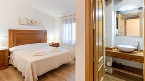Un dormitorio con cabecera de madera, una mesita de noche con lámpara, cama con ropa blanca de cama y un baño con tocador de madera y lavamanos.