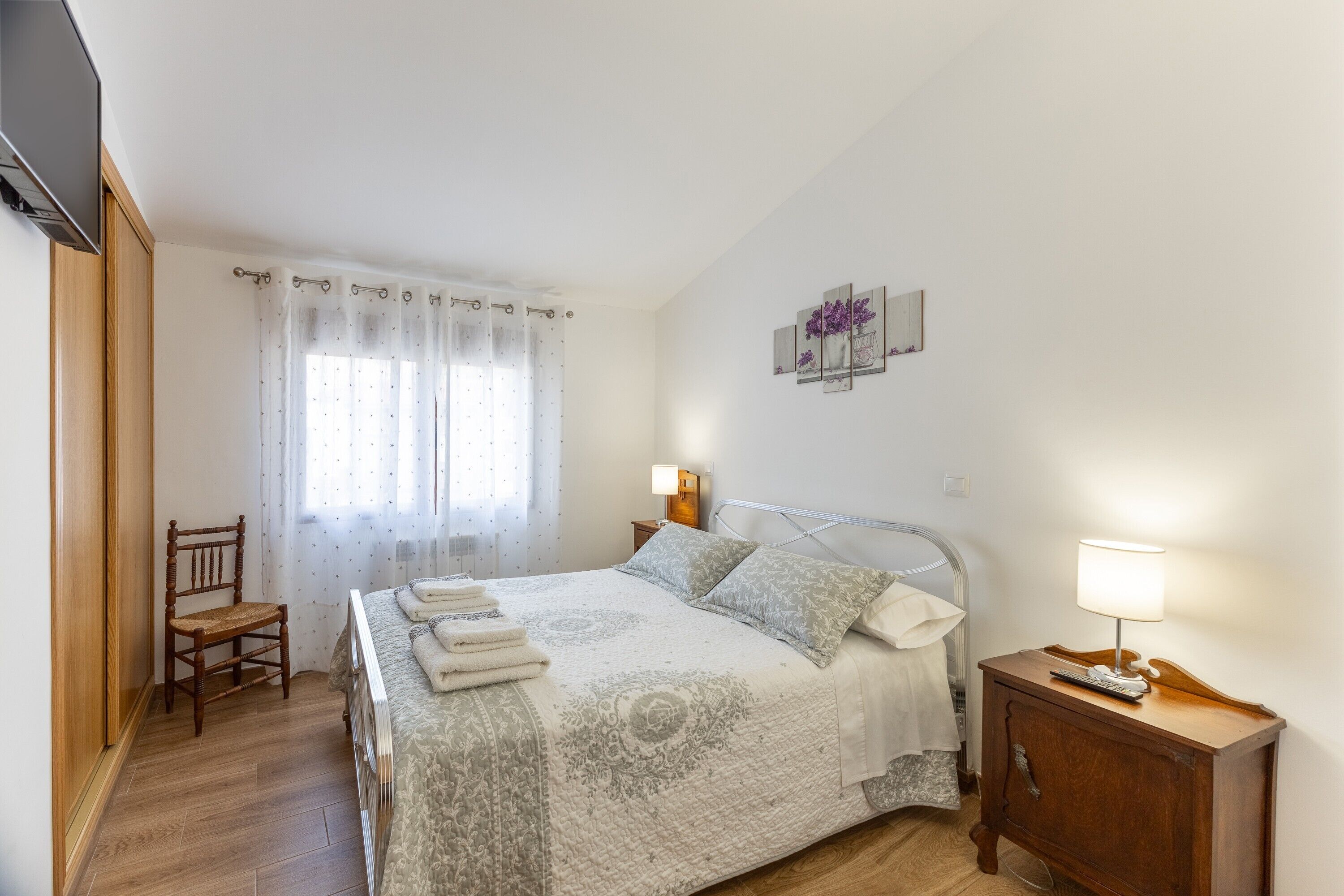 Un dormitorio con cama, una silla, una mesita de noche, una lámpara y una obra de arte en la pared.