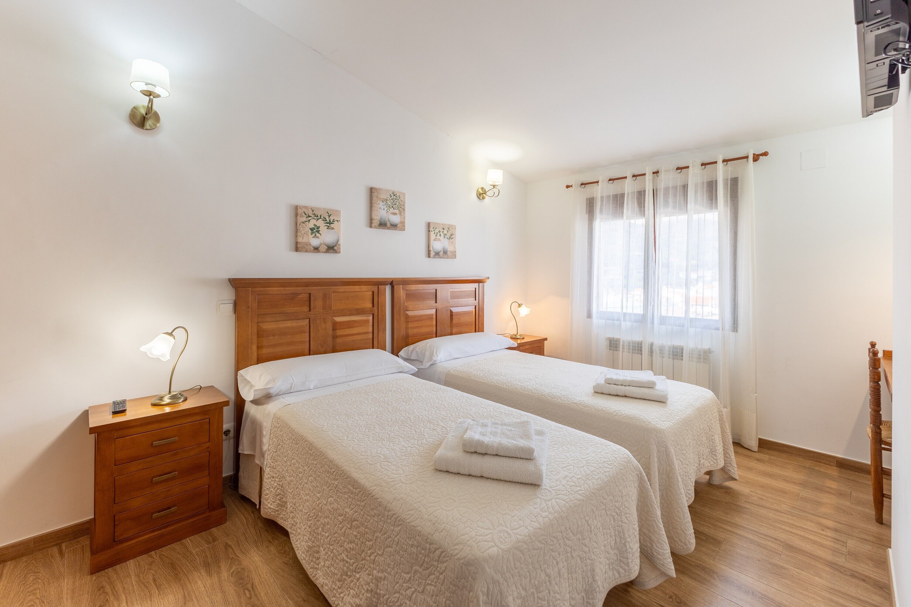 Habitación doble con dos camas, cabecero de madera, mesitas de noche con lámparas, ventana con cortinas traslúcidas y una obra de arte colgada en la pared.