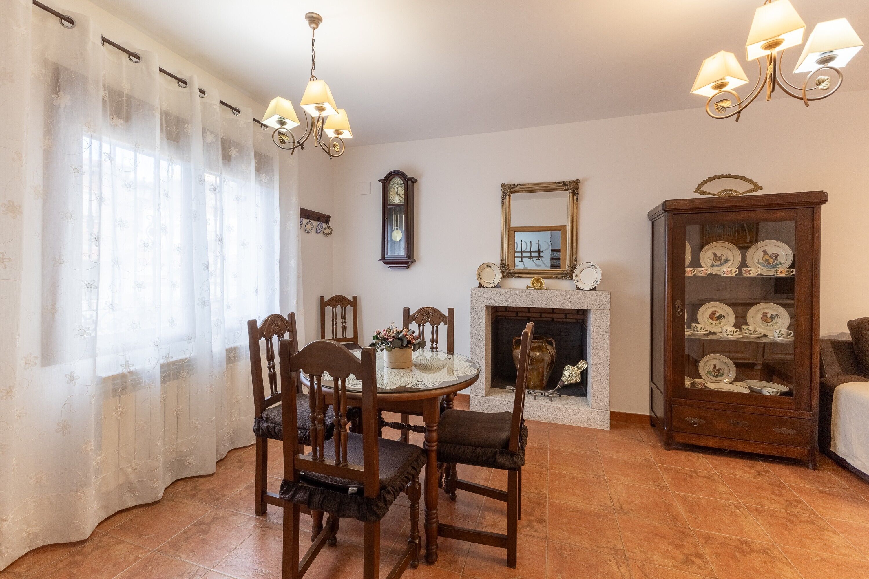 A dining area with a round table, chairs, a chandelier, a fireplace, a clock, and a china cabinet.