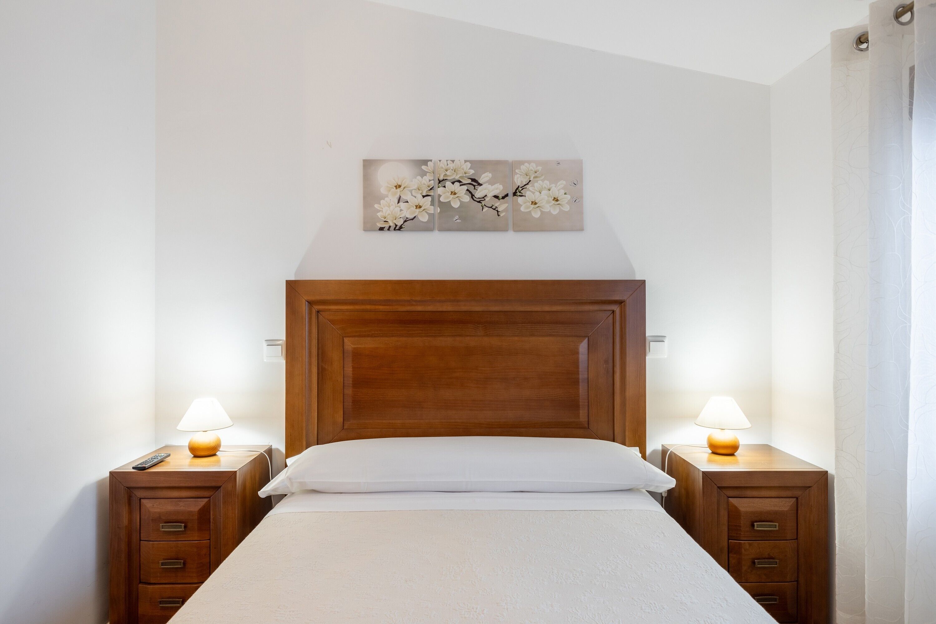 Een netjes opgemaakt bed met een houten hoofdbord, twee nachtkastjes met lampen en een kunstwerk aan de muur erboven.