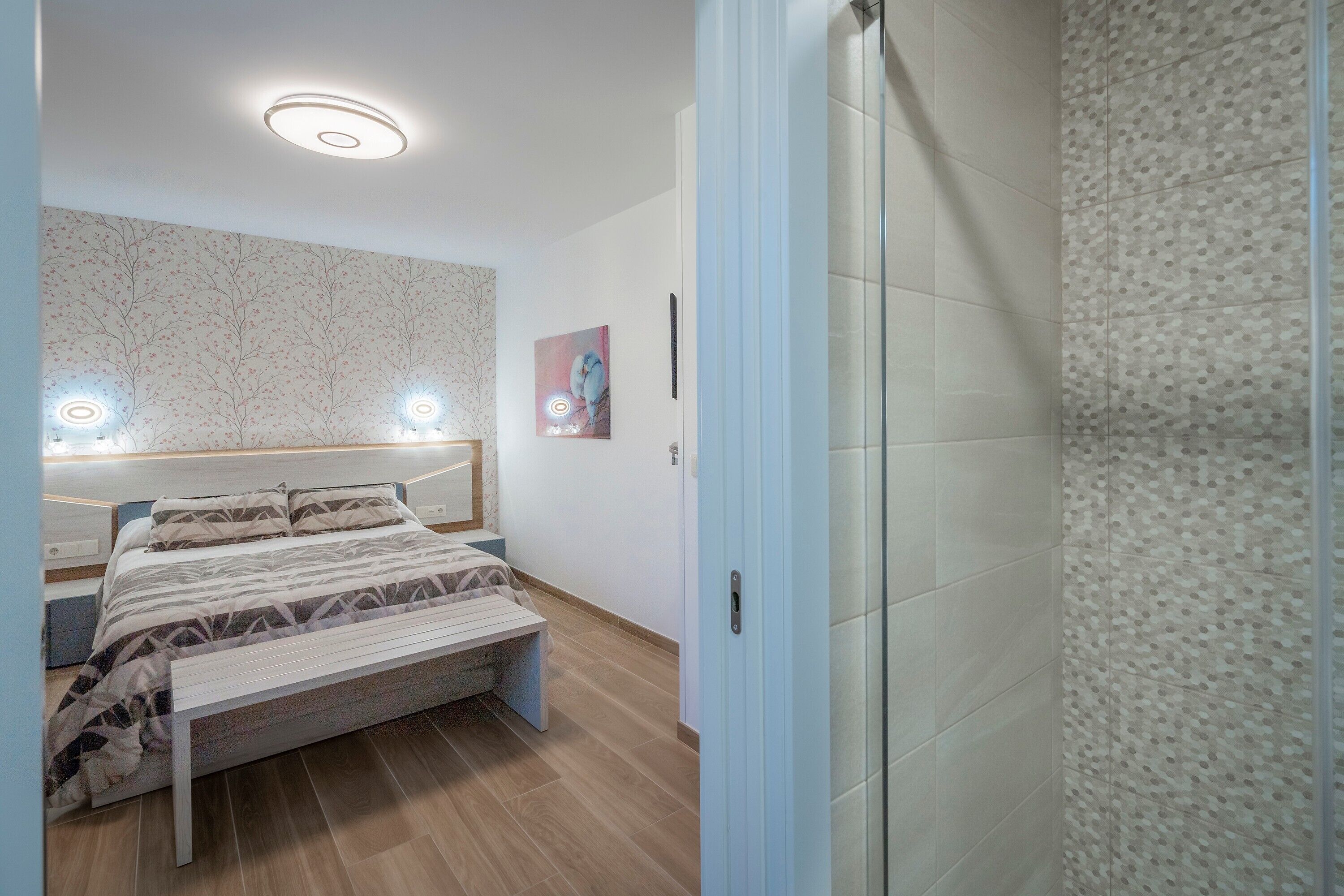 A bedroom with a bed, bedside tables, a bench, a wall with a tree pattern, and a door leading to another room.
