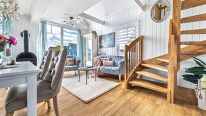 A spacious living room with a wooden staircase, a fireplace, and large windows. - La petite maison bleue (Wandlitz OT Zerpenschleuse)
