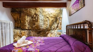 Un lit avec une couverture violette, une tête de lit en bois et un mur de pierre derrière.
