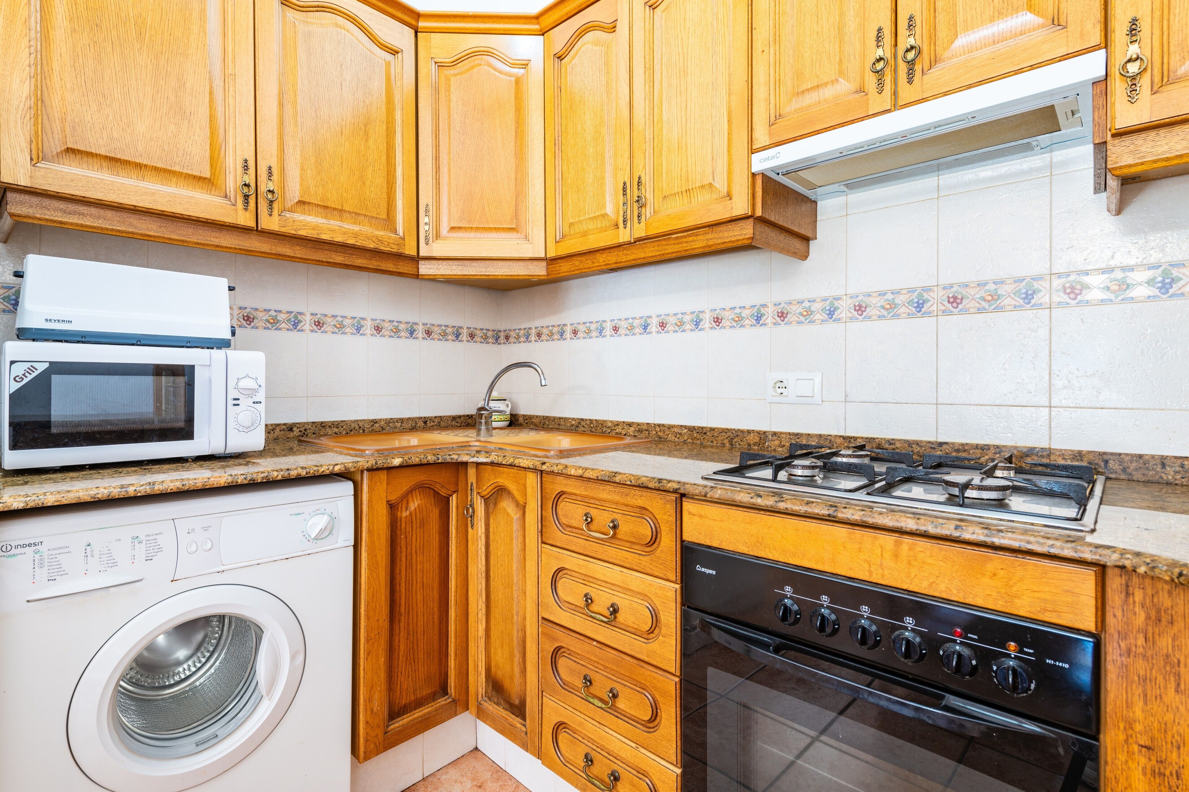 Een keuken met houten kastjes, een magnetron, een wasmachine, een spoelbak en een fornuis.