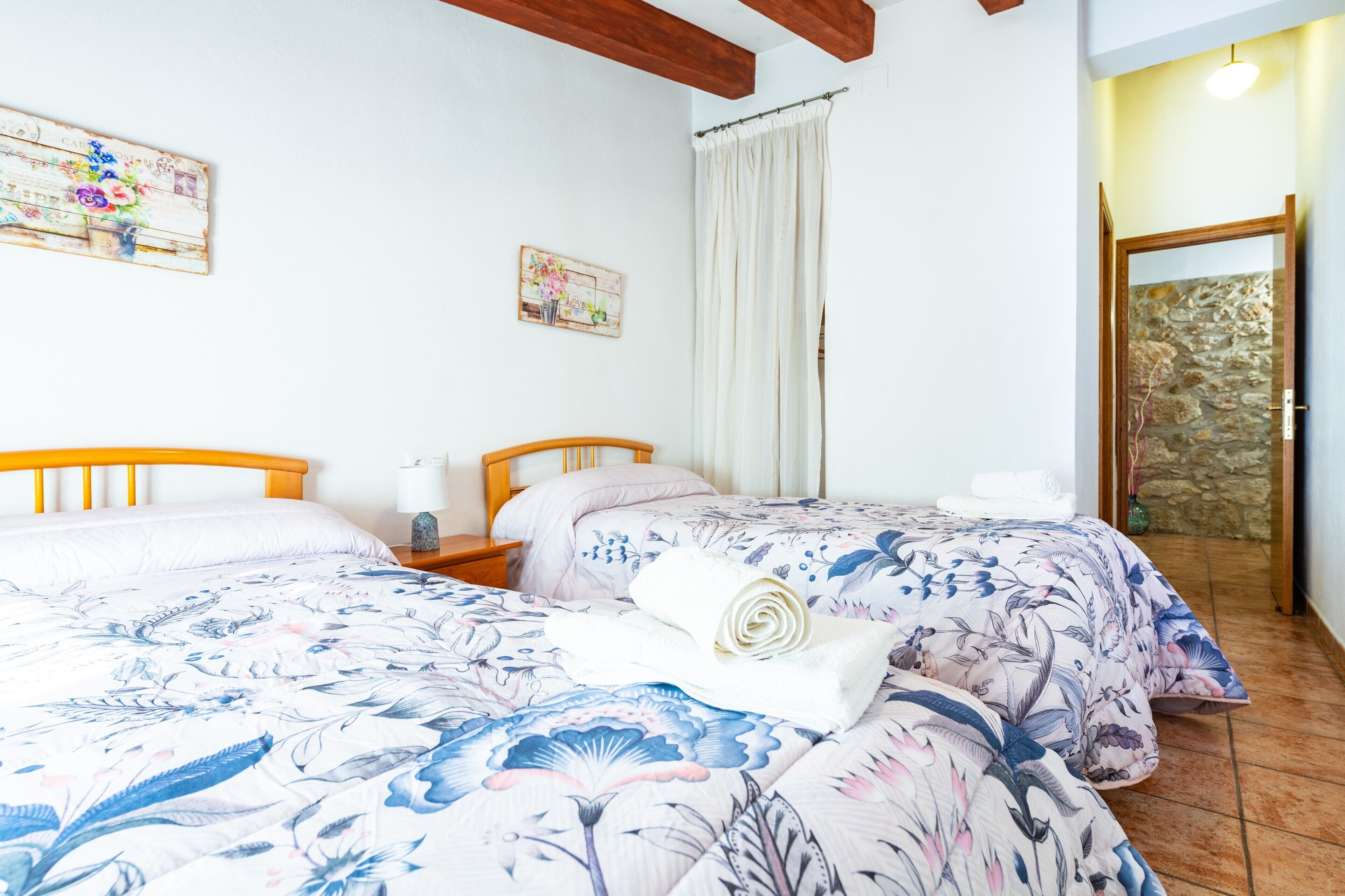 Une chambre avec deux lits, une tête de lit en bois, une table de chevet, une serviette blanche, un tableau, un rideau et un mur en pierre.