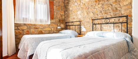 A bedroom with two beds, stone walls, wooden beams, and a window with curtains.