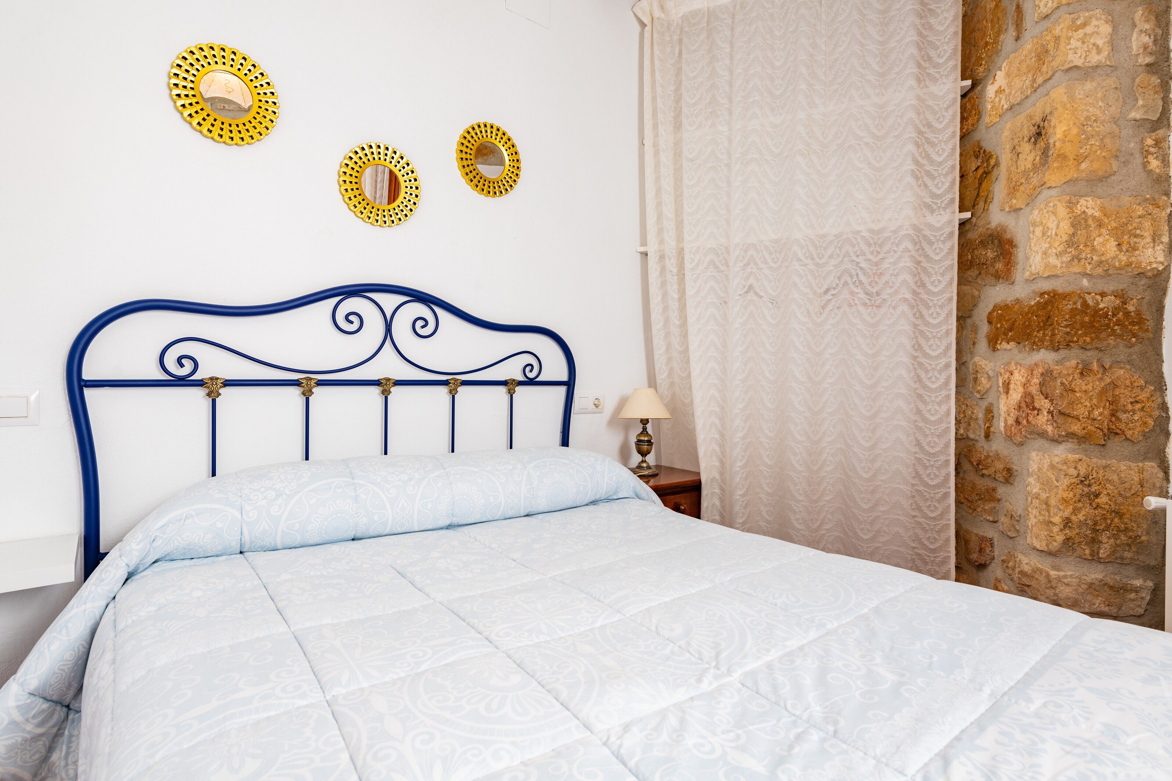 A bed with a blue headboard, a white wall with three circular mirrors, and a stone wall.