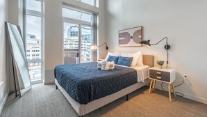 A modern bedroom with a large bed, bedside tables, and a view of the city through large windows. - Sophari Downtown: Bellevue Apartments (Bellevue)
