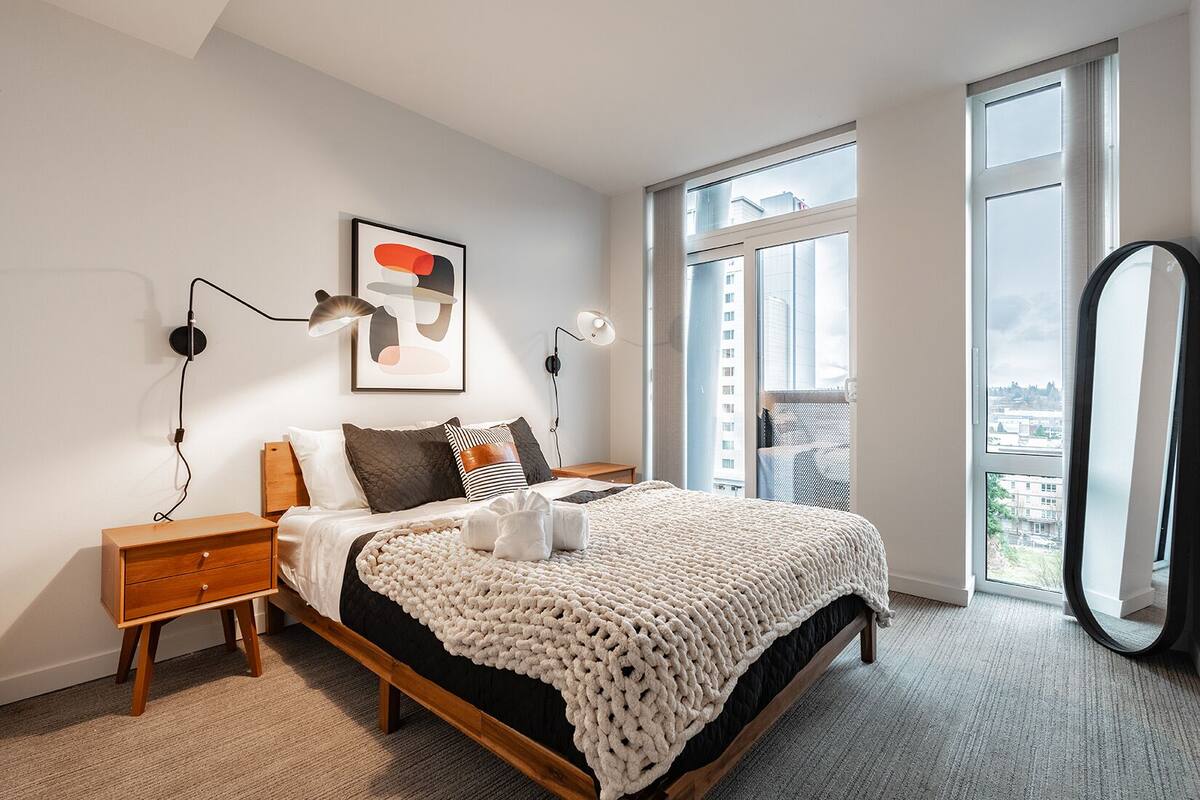 A modern bedroom with a large bed, a nightstand, a wall-mounted lamp, a framed abstract artwork, and a large window with a view of buildings.