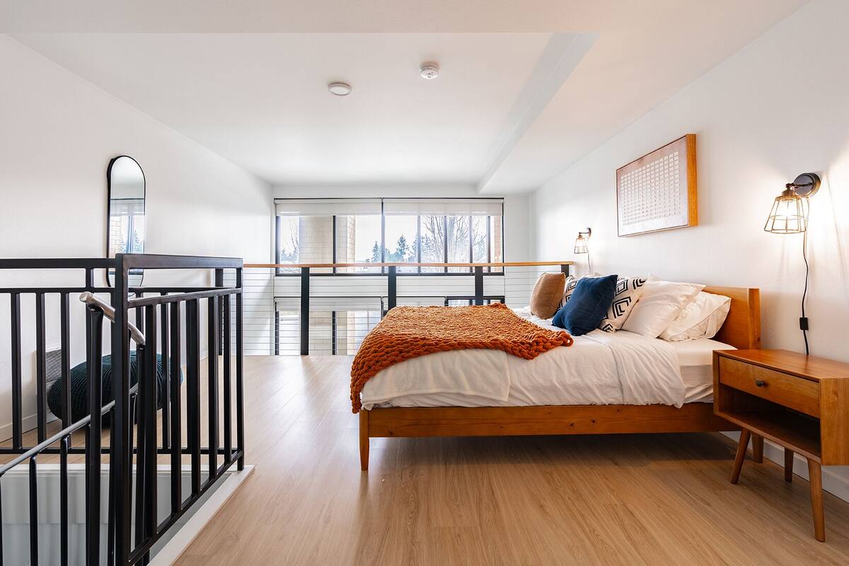 A modern bedroom with a wooden bed, a nightstand, a window with a view, a balcony, and a staircase.