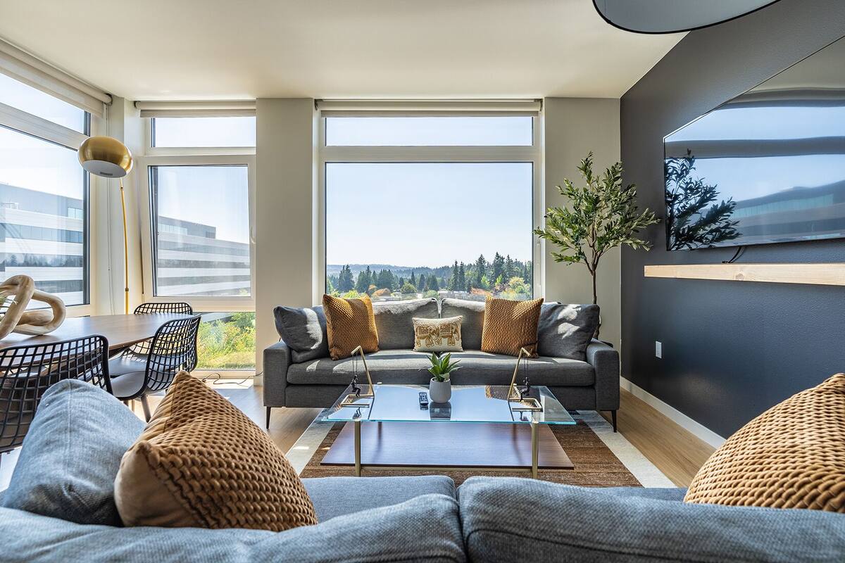 A modern living room with a grey sofa, a glass coffee table, and a view of a forest through large windows.