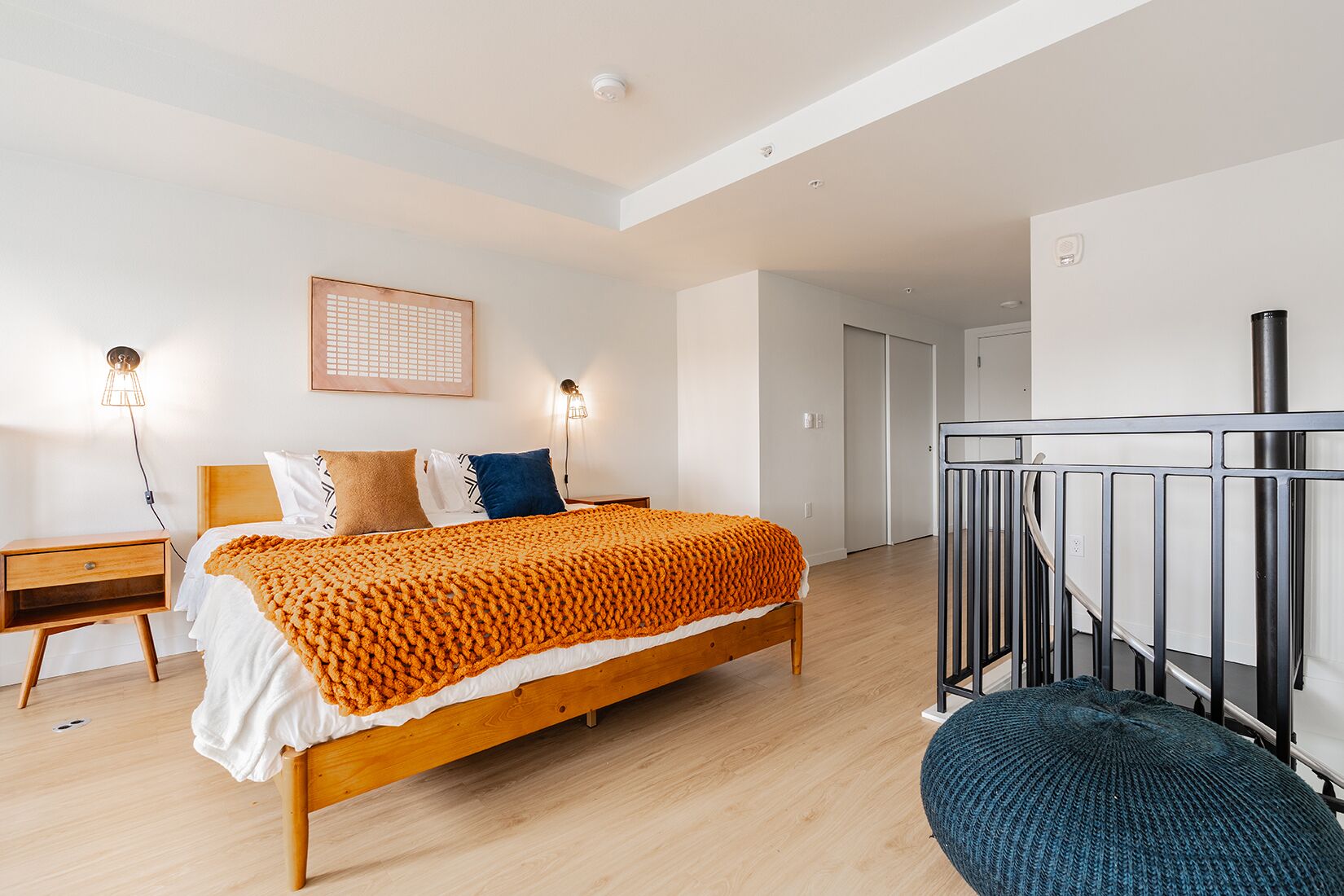 A modern bedroom with a wooden bed, a nightstand, a wall-mounted lamp, and a staircase with a railing.
