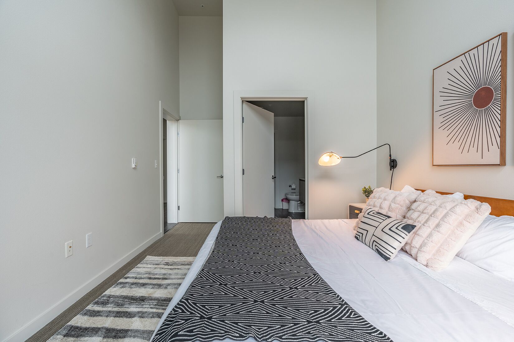 Un dormitorio moderno con una cama grande, un tapete estampado, una mesita de noche, una lámpara de pared y una puerta que da al baño.