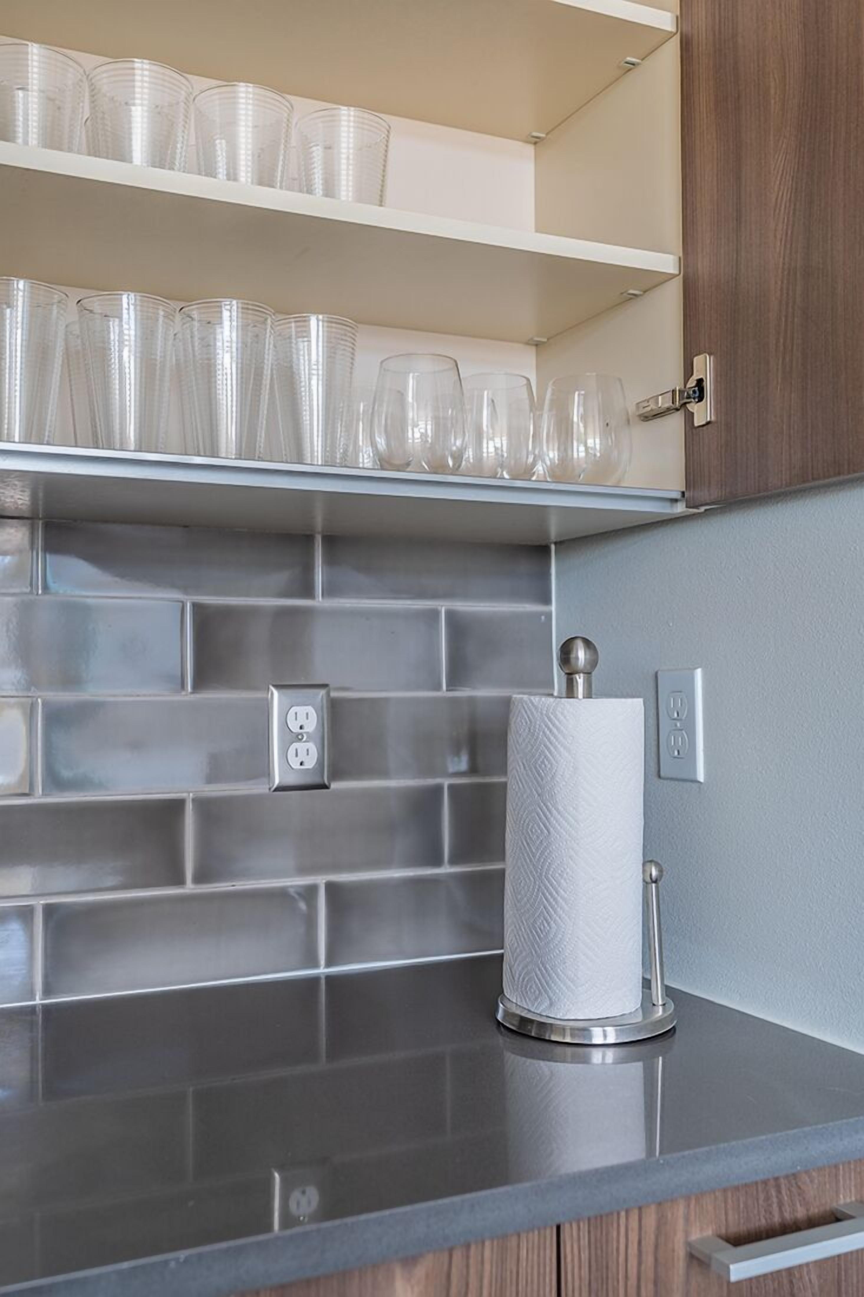 A kitchen with a countertop, a paper towel holder, and a shelf with clear plastic cups and glasses.