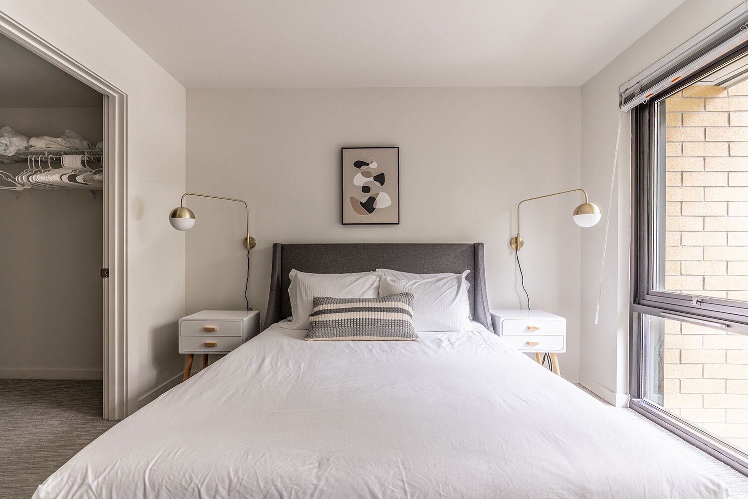 A neatly arranged bedroom with a large bed, bedside tables, a framed artwork above the bed, and a window providing natural light.