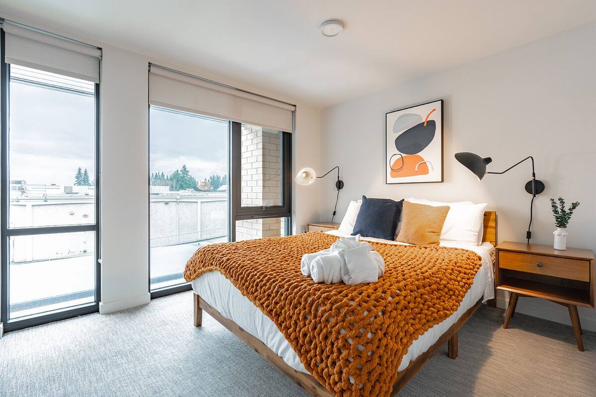 A modern bedroom with a large bed, a nightstand, a floor lamp, and a view of the city through large windows.
