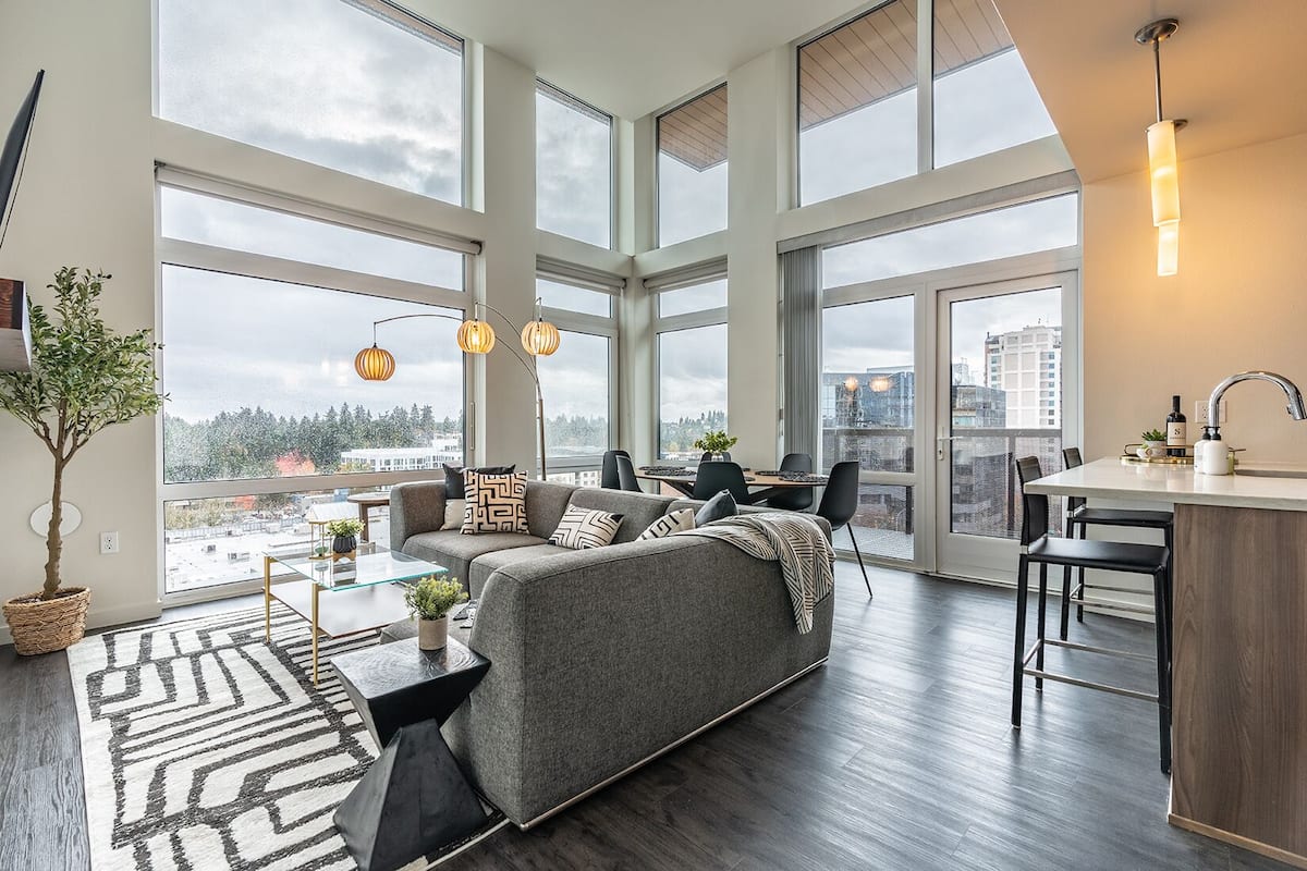 A modern apartment with a large living area, dining space, and kitchen. It features large windows, a grey sofa, and a dining table with chairs.