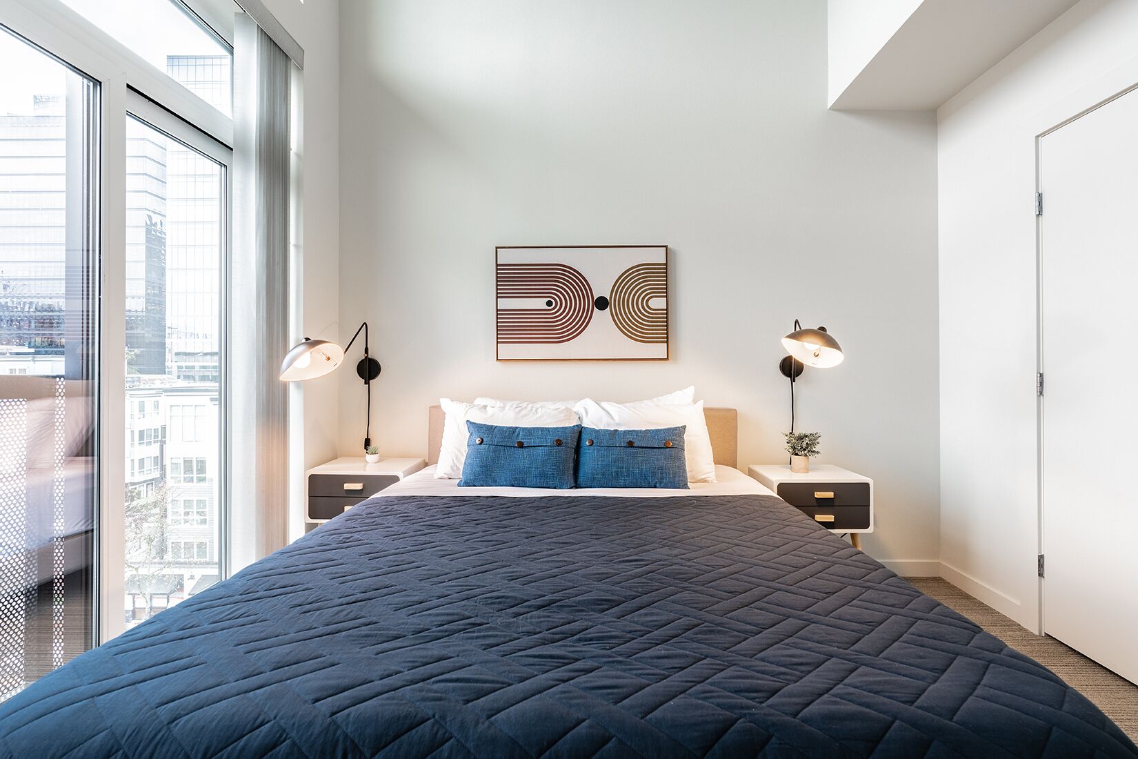 Um quarto moderno com uma cama grande, duas mesas de cabeceira, uma obra de arte na parede e vista para a cidade através de amplas janelas.