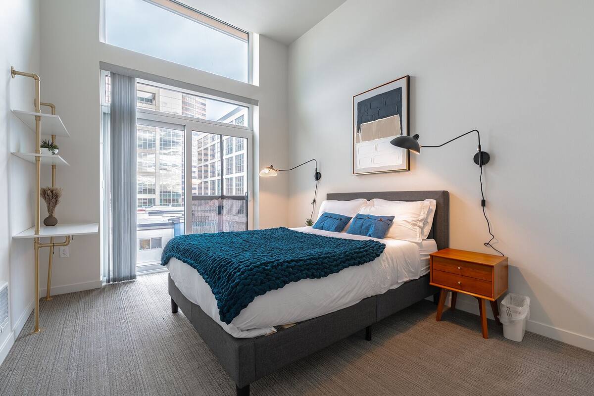 A modern bedroom with a large window, a bed with a blue blanket, a bedside table, and a wall-mounted lamp.