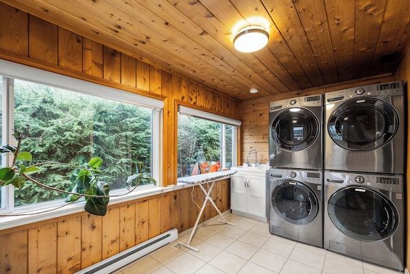 A laundry room with wooden walls, large windows, and four stacked front-loading washing machines. - Oceanfront Cottage: Dock, Beach Access, Hot Tub, BBQ & Free Kayaks (Sooke)