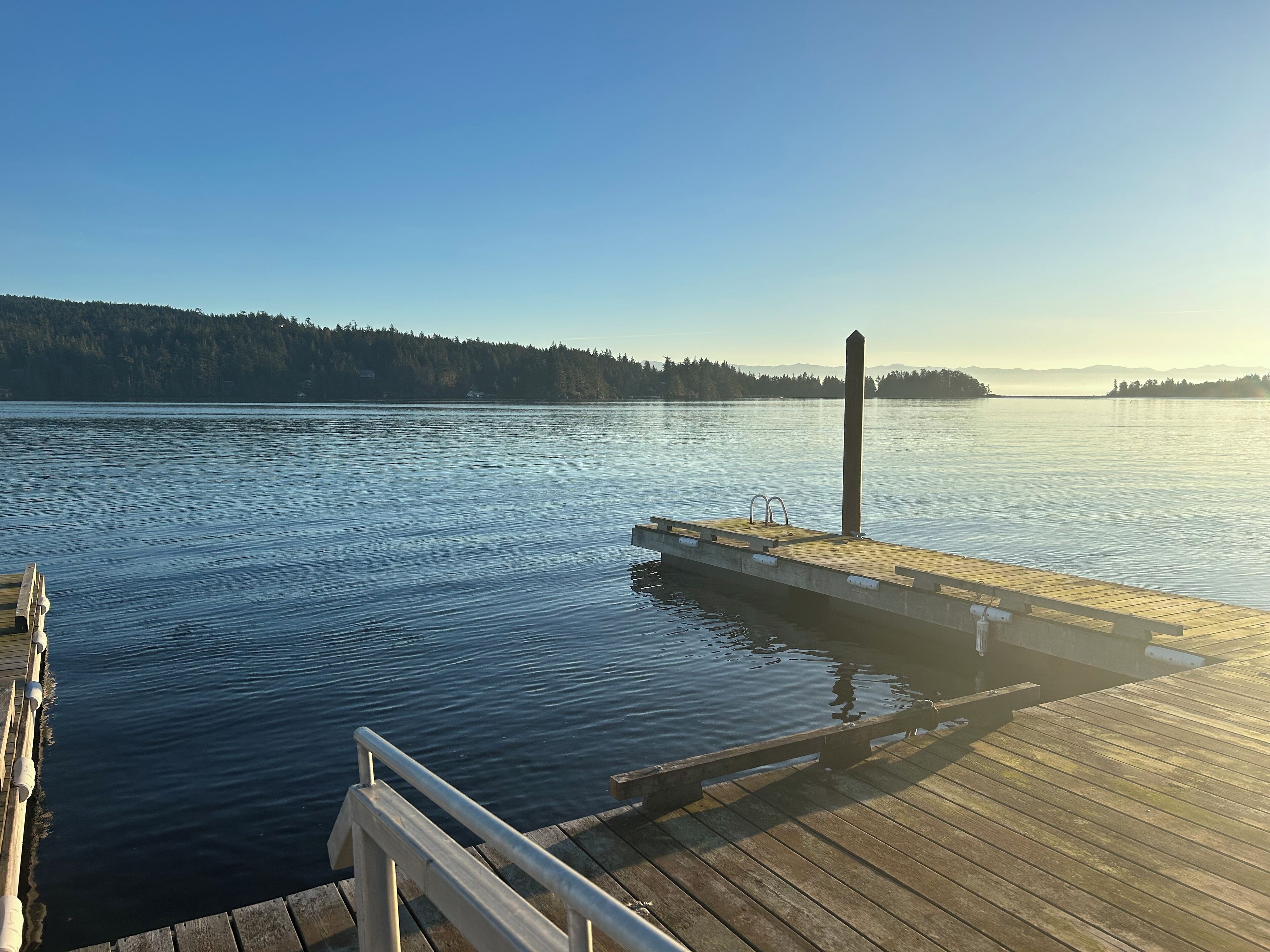 A wooden dock extending into calm water with a clear sky and forested hills in the background.