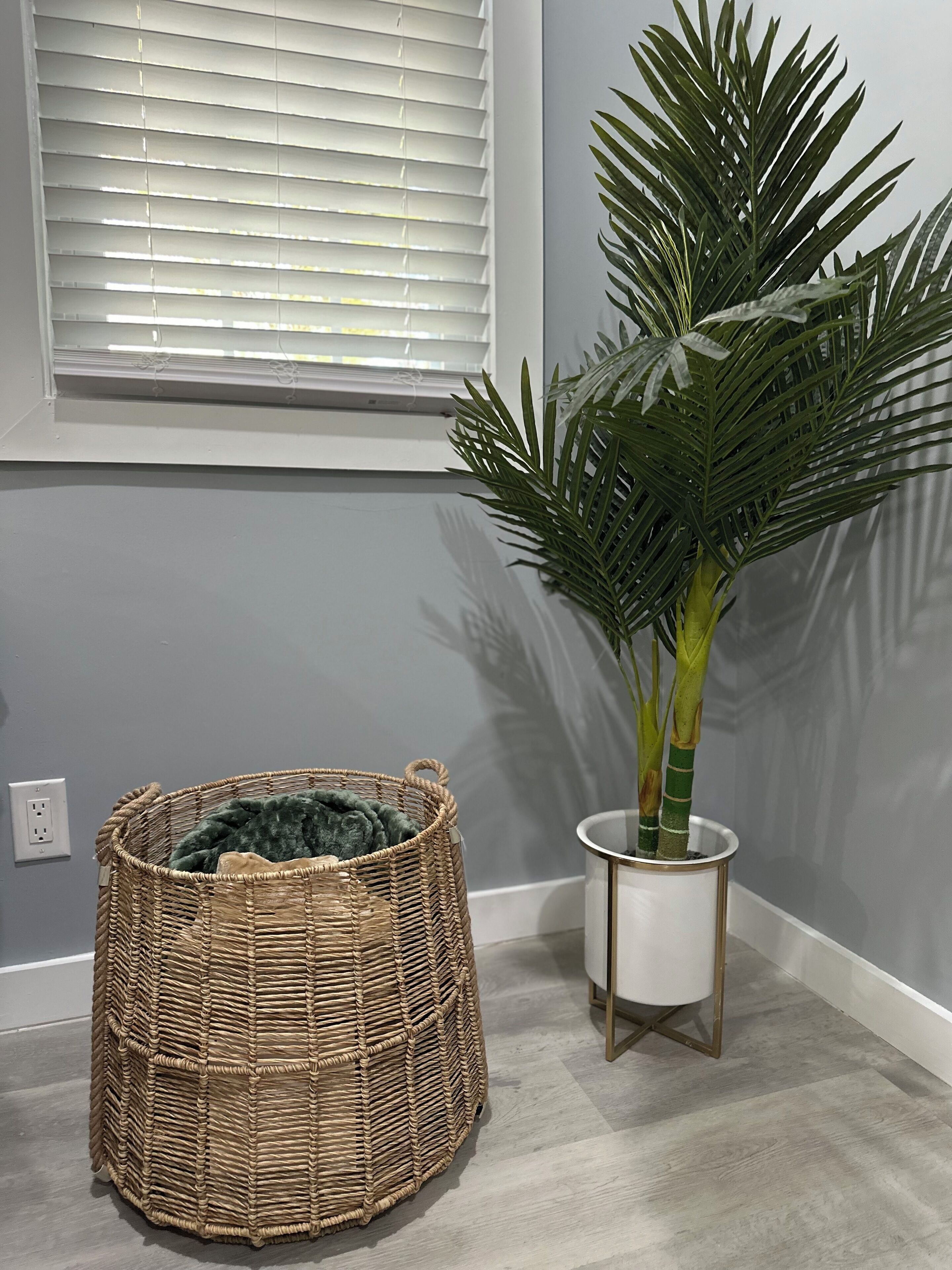 A corner of a room with a woven basket, a potted plant, and a window with blinds.