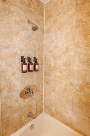Une salle de bain avec des carreaux de marbre beige, une douche, une baignoire et trois bouteilles de shampoing.