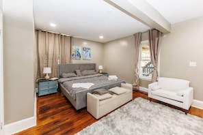 A bedroom with a bed, a nightstand, a dresser, a white armchair, and a window with curtains.