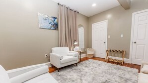 Ein Zimmer mit einem weißen Sofa, einem kleinen Tisch mit einer Lampe, einem grauen Shaggy-Teppich und einem Holzregal.