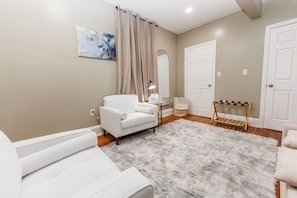 A room with a white sofa, a small table with a lamp, a grey shag rug, and a wooden rack.
