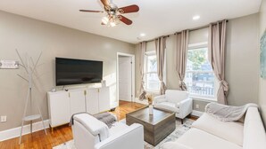 Ein modernes Wohnzimmer mit Flachbildfernseher, weißen Sofas, einem Couchtisch und einem Deckenventilator.