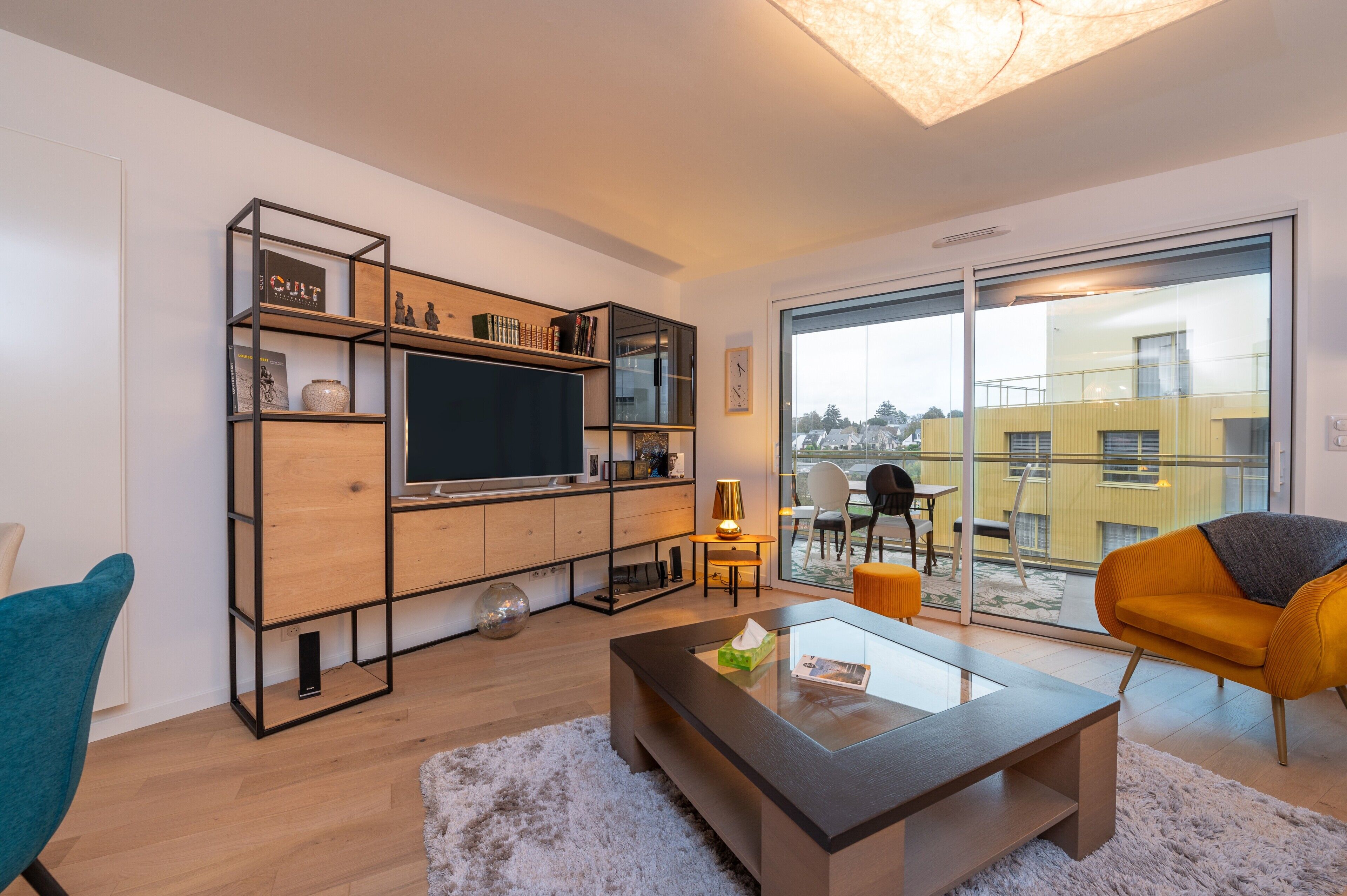 A modern living room with a flat-screen TV, wooden shelving, a glass coffee table, and a view of a yellow building through large windows.