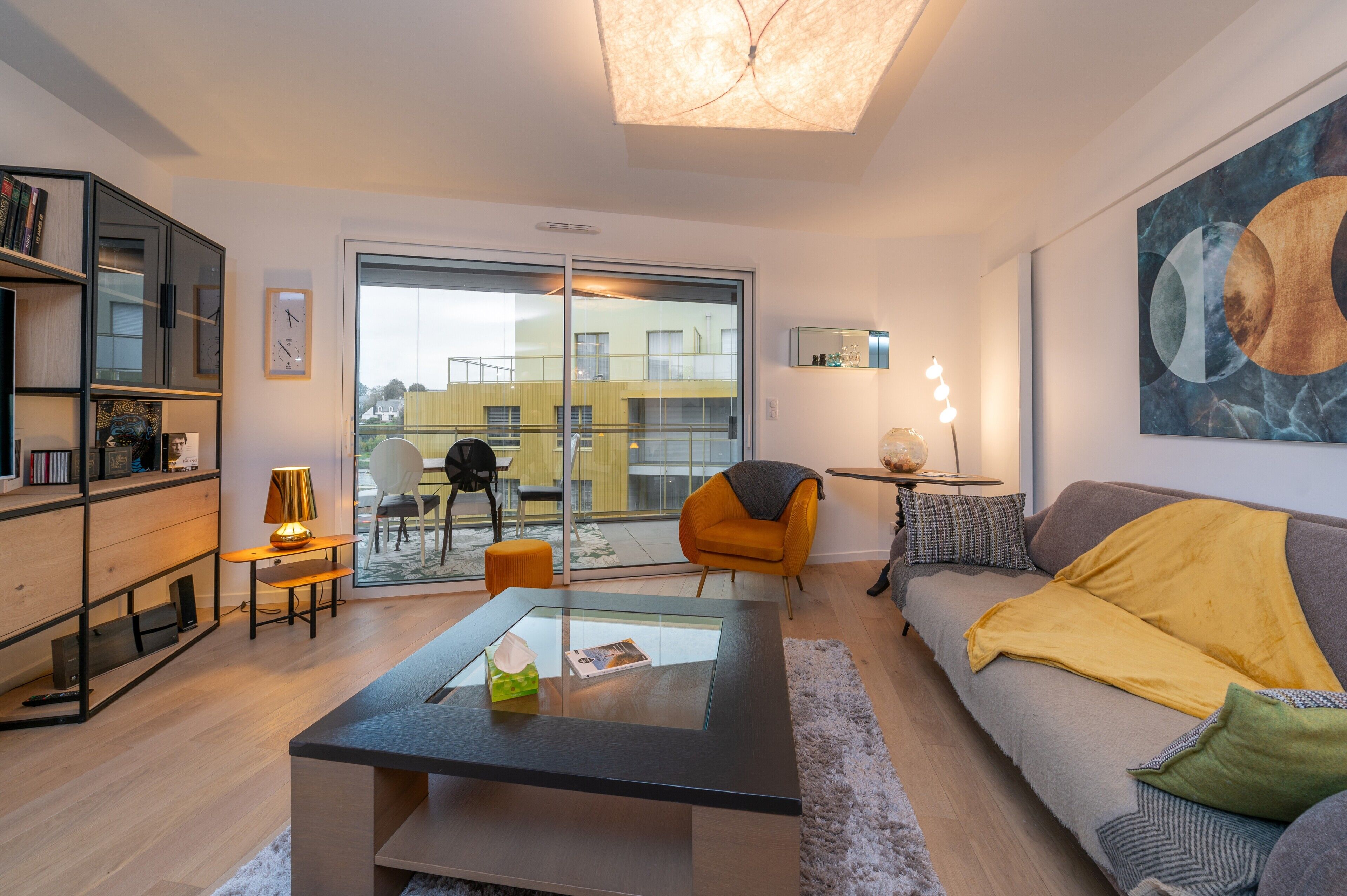 Una sala moderna con una mesa de centro de vidrio, un sofá con una manta amarilla, una estantería, una silla y un ventanal con vistas a edificios.