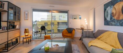 Una sala moderna con una mesa de centro de vidrio, un sofá con una manta amarilla, una estantería, una silla y un ventanal con vistas a edificios.