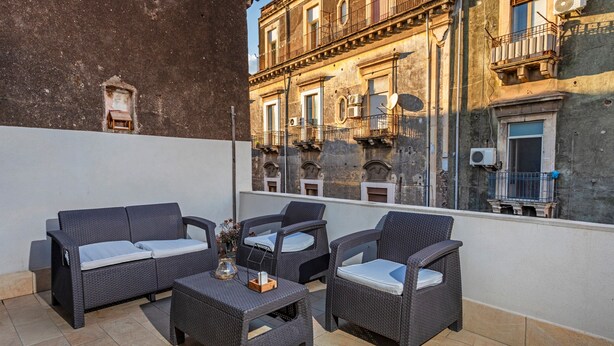 Un balcón con mobiliario de mimbre, una pequeña mesa y vistas a un edificio con balcones y unidades de aire acondicionado.