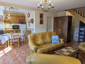 Uma sala de estar com sofá, mesa de centro e uma área de jantar com cadeiras e mesa. Cozinha visível ao fundo, com armários e eletrodomésticos.