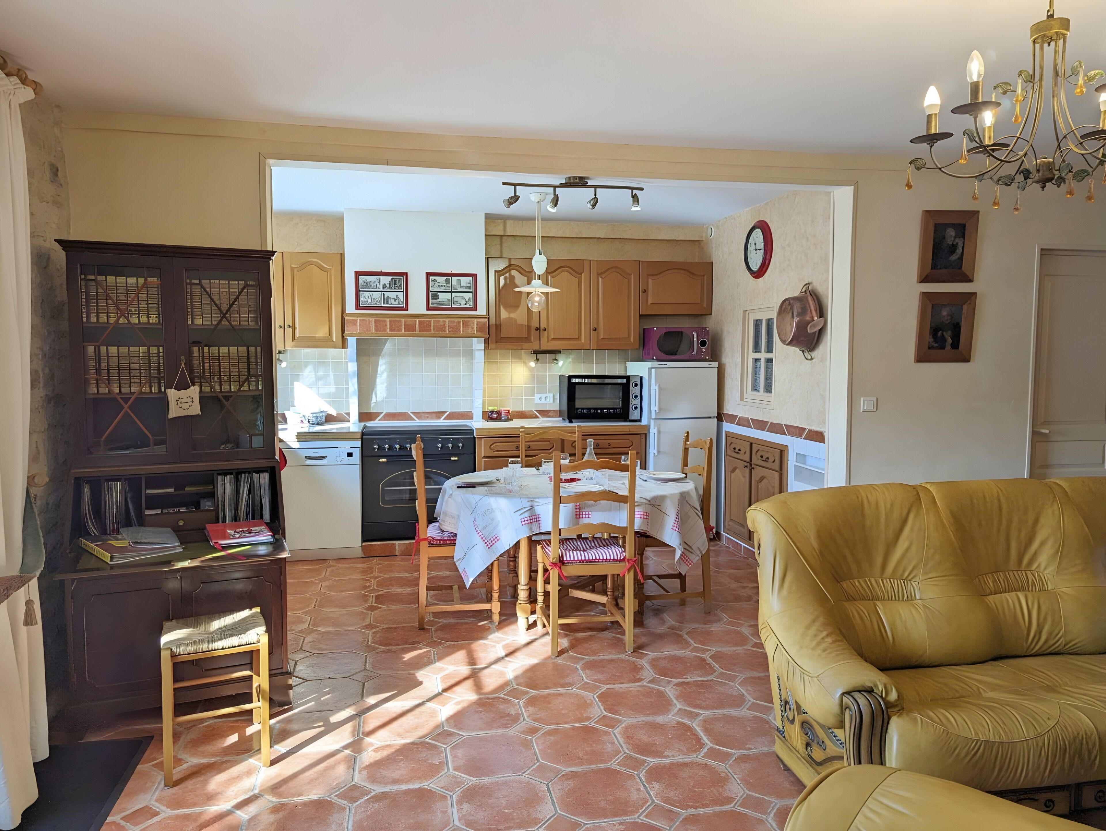 A spacious living area with a sofa, a dining table with chairs, a kitchen with wooden cabinets and appliances, and a chandelier.