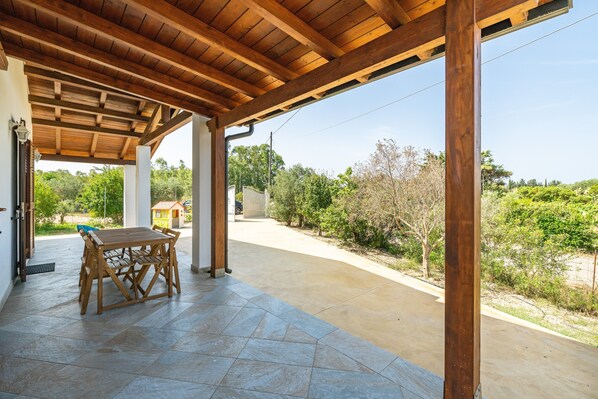 Un patio cubierto con una mesa y sillas de madera, piso de baldosas y vistas a la vegetación y un edificio a lo lejos.