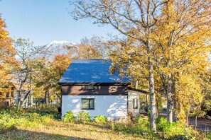 A two-story house with a blue roof, surrounded by trees with autumn foliage, and a snow-capped mountain in the background. - Gensei (Kutchan)