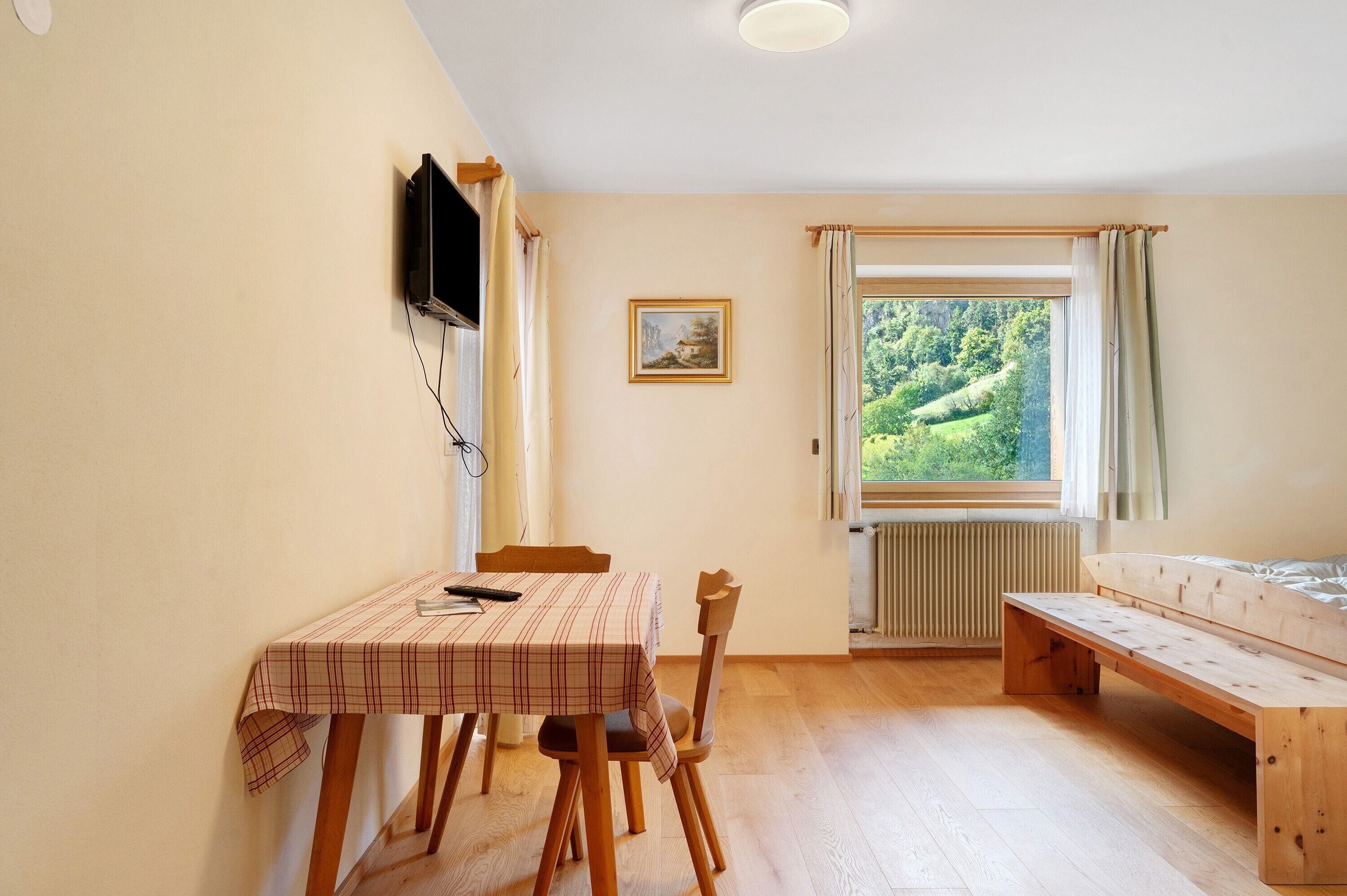 Ett rum med ett bord, stolar, en bänk, en tv som är monterad på väggen, ett fönster med gardiner och ett element.