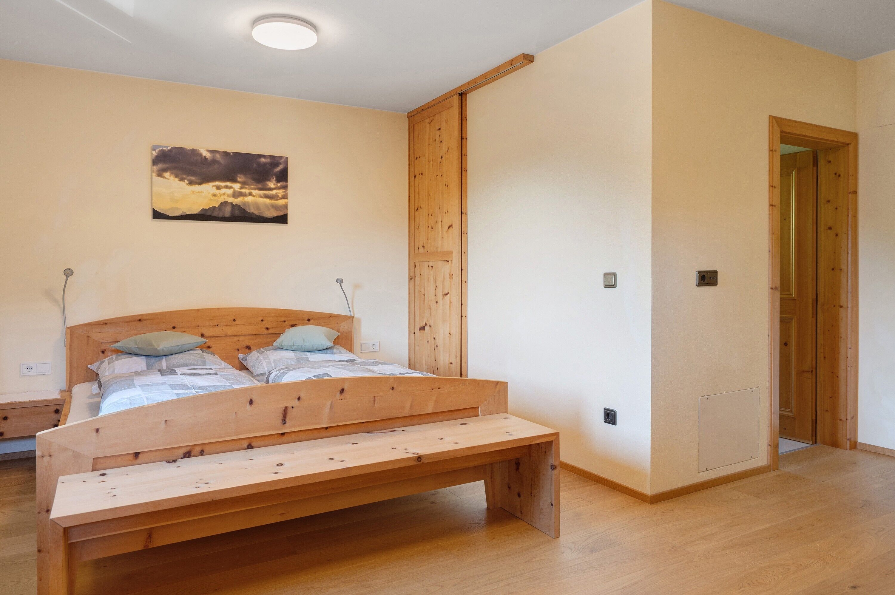 Un dormitorio con una cama de madera, un banco, un cuadro en la pared y una puerta que da a otra habitación.