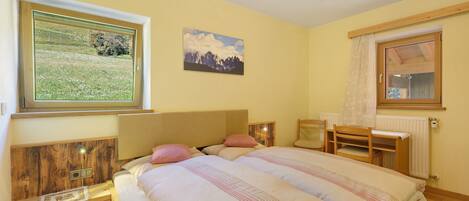 Une chambre avec deux lits, une fenêtre donnant sur un champ, un tableau représentant des montagnes et une tête de lit en bois.