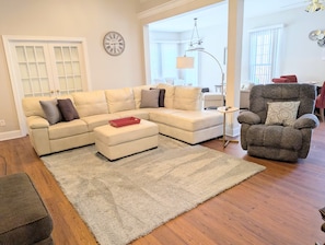 A living room with a sectional sofa, a small ottoman, a grey rug, a grey armchair, and a floor lamp.