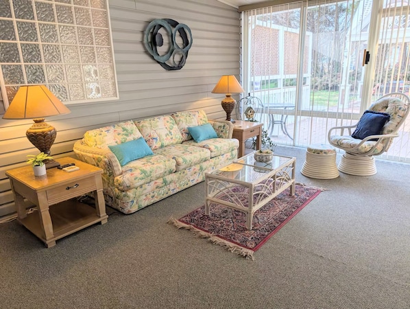 A cozy living room with a floral sofa, two side tables with lamps, a glass coffee table, a wicker chair, and a view through a large window.